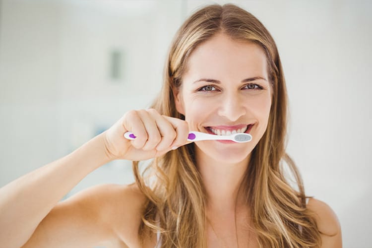 Woman brushing teeth in bathroom, smiling with white teeth.