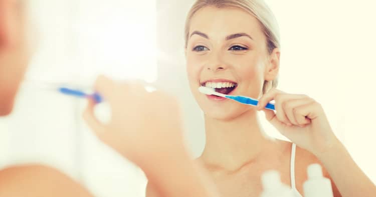 a woman is brushing her teeth in front of a mirror wondering "How often should I replace my toothbrush?"