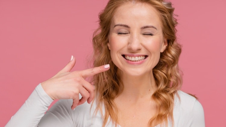 A young woman who has received composite veneers, smiling confidently and pointing at her enhanced teeth to show the results.