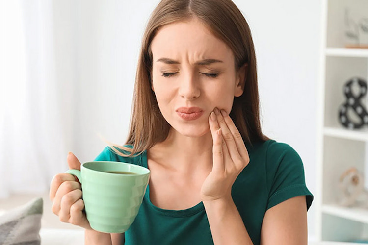 a woman is holding her hand on her cheek while drinking tea, showing she's suffering from dental pain.